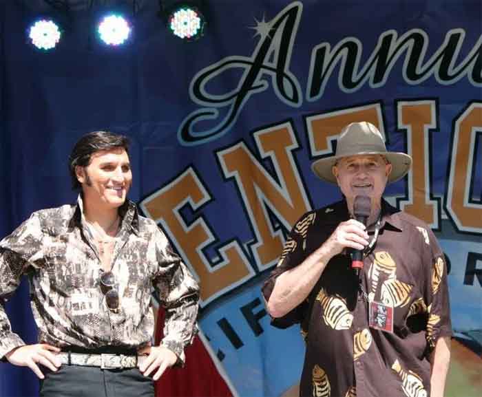 Two men smiling on stage at an event with a microphone and banner behind them.